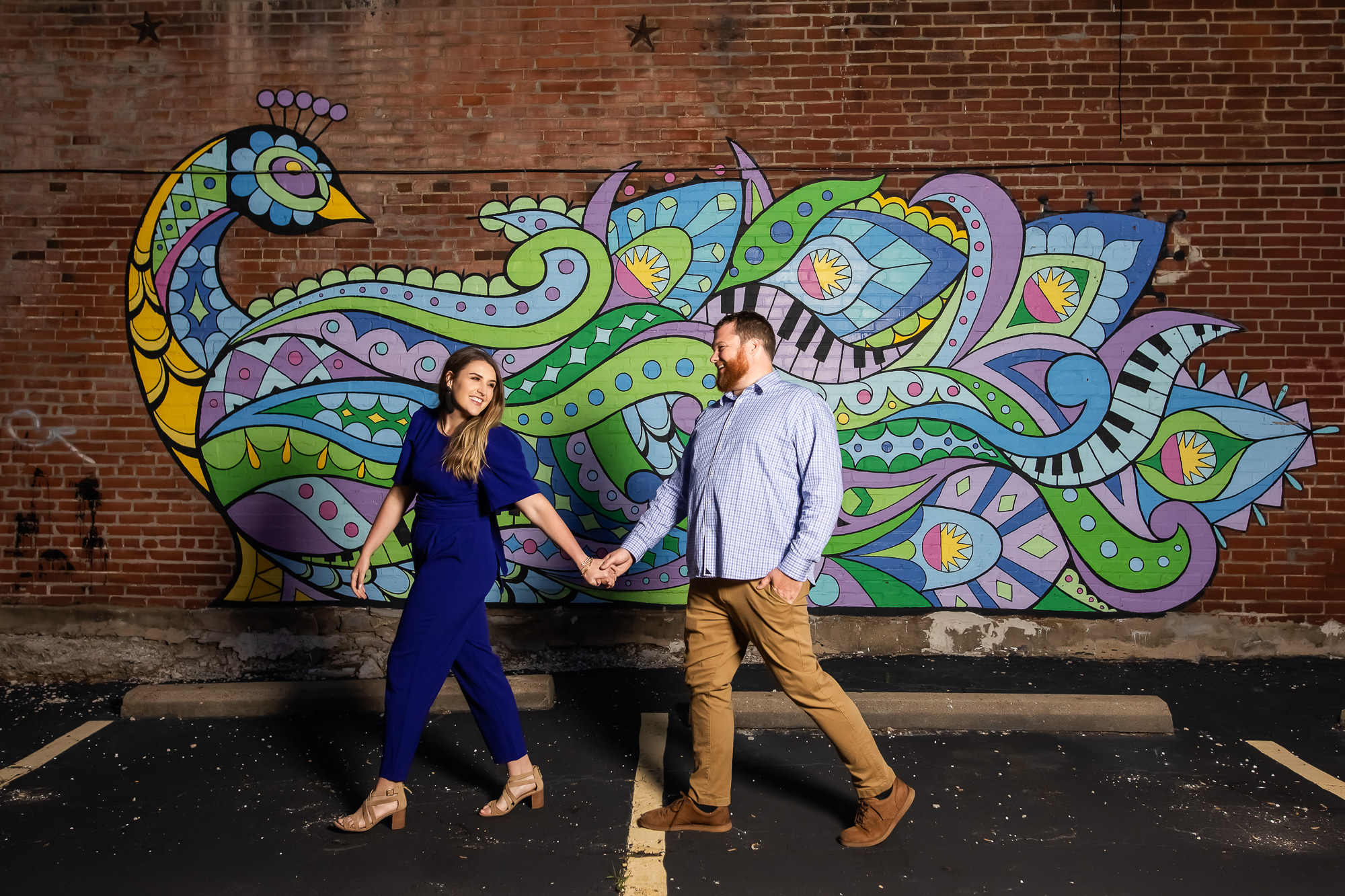 Engaged couple holding hands and walking in front of a brick wall with a peacock mural