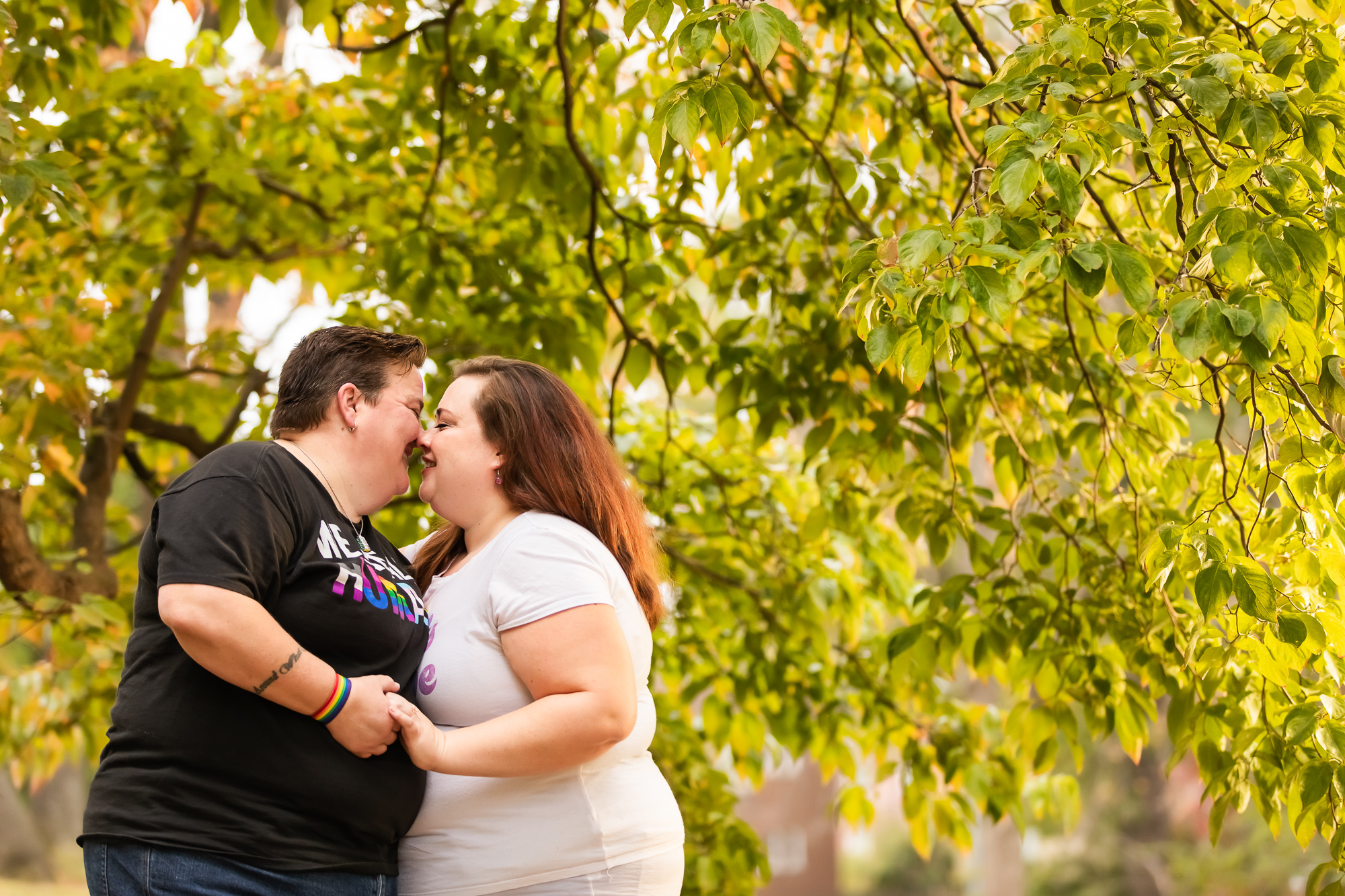 Lesbian couple holding hands and kissing beneath a tree.